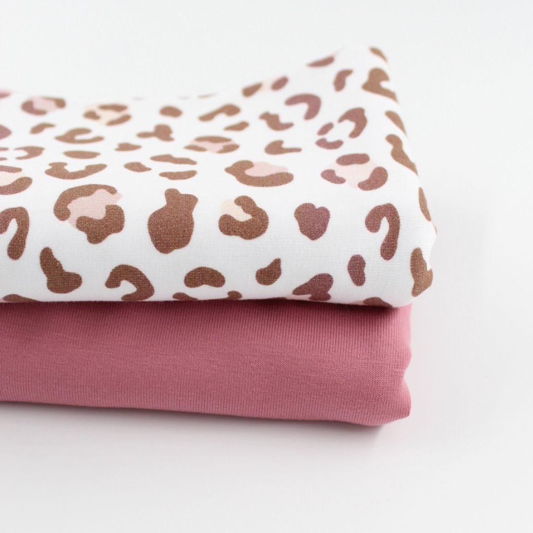 Zwei aufeinandergelegte Jersey-Stoffe. Der obere Stoff hat ein modernes Leopardenmuster in Braun- und Rosatönen auf weißem Hintergrund. Der untere Stoff ist ein einfarbiger, zart rosafarbener Jersey. Beide Stoffe eignen sich perfekt für Kleidung und Accessoires.