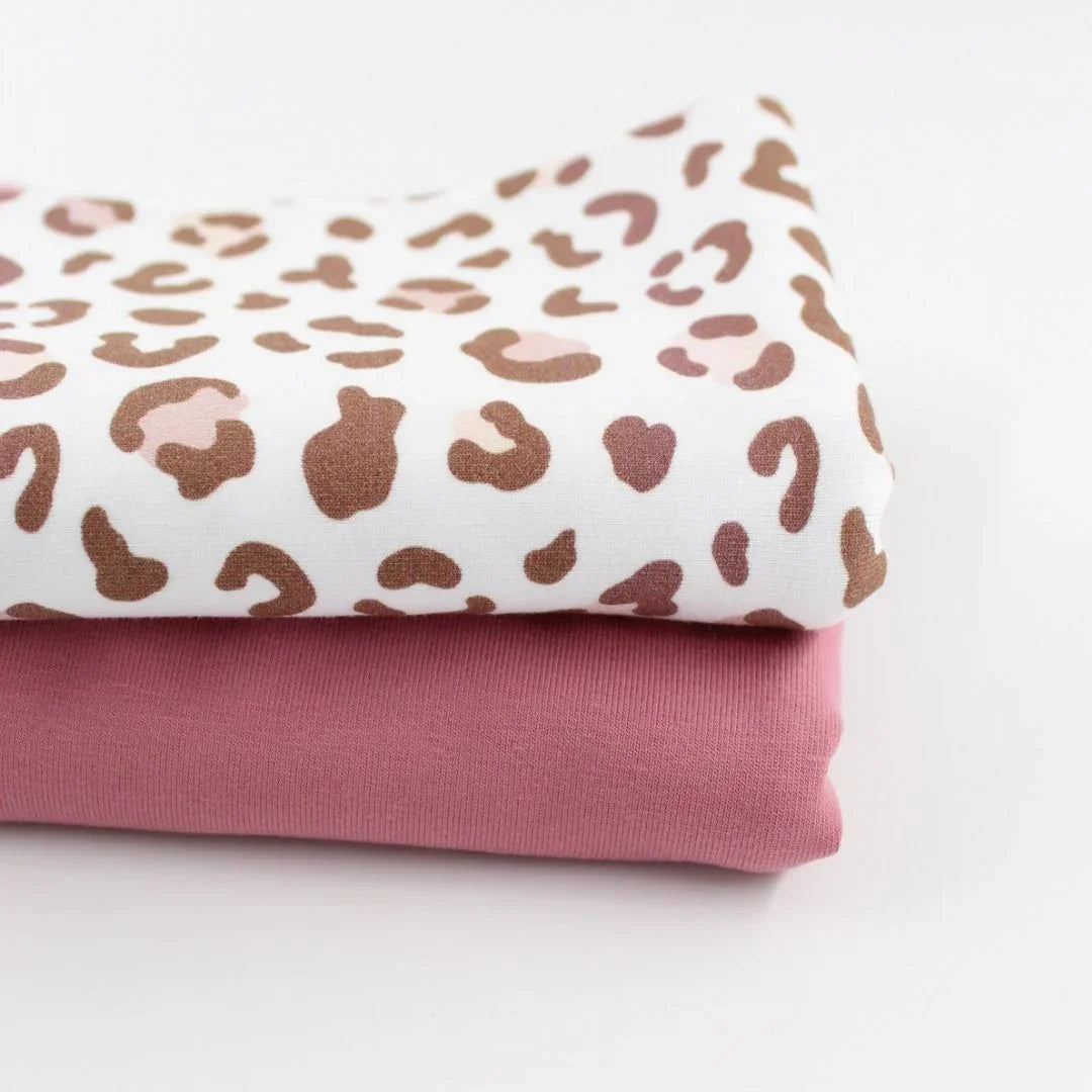 Zwei aufeinandergelegte Jersey-Stoffe. Der obere Stoff hat ein modernes Leopardenmuster in Braun- und Rosatönen auf weißem Hintergrund. Der untere Stoff ist ein einfarbiger, zart rosafarbener Jersey. Beide Stoffe eignen sich perfekt für Kleidung und Accessoires.