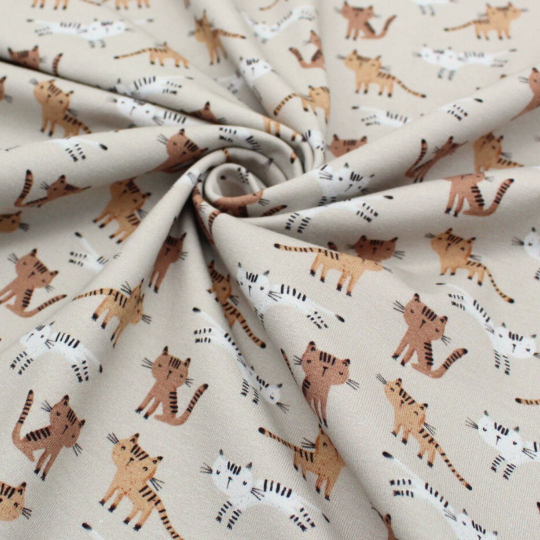 Der Jersey-Stoff mit niedlichem Katzenmuster auf beigem Hintergrund, perfekt für Nähprojekte wie T-Shirts und Kleider.