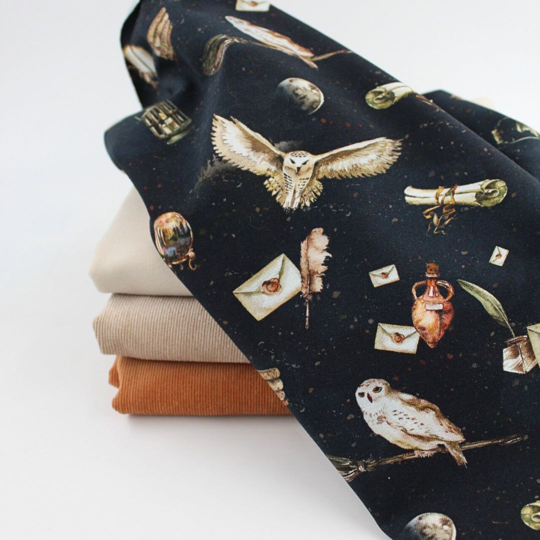 Stoffe zum Nähen: Oben Jersey mit magischen Motiven wie Eulen, Briefe, Schriftrollen und Zaubertränke auf schwarzem Hintergrund. Darunter French Terry in Sand sowie Feincord in Sand und Karamellbraun gestapelt.