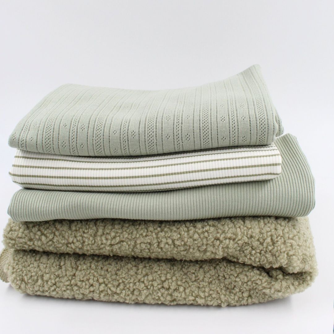 Ein Stapel sorgfältig gefalteter Stoffe in sanften Grüntönen, bestehend aus verschiedenen Texturen: ein zart gestrickter Stoff mit Lochmuster oben, darunter zwei gestreifte Jerseys, ein gerippter Stoff und unten ein weicher, flauschiger Bouclé-Stoff. Die neutrale Farbpalette und die unterschiedlichen Materialien verleihen dem Bild eine harmonische und gemütliche Ausstrahlung.