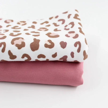 Zwei aufeinandergelegte Jersey-Stoffe. Der obere Stoff hat ein modernes Leopardenmuster in Braun- und Rosatönen auf weißem Hintergrund. Der untere Stoff ist ein einfarbiger, zart rosafarbener Jersey. Beide Stoffe eignen sich perfekt für Kleidung und Accessoires.