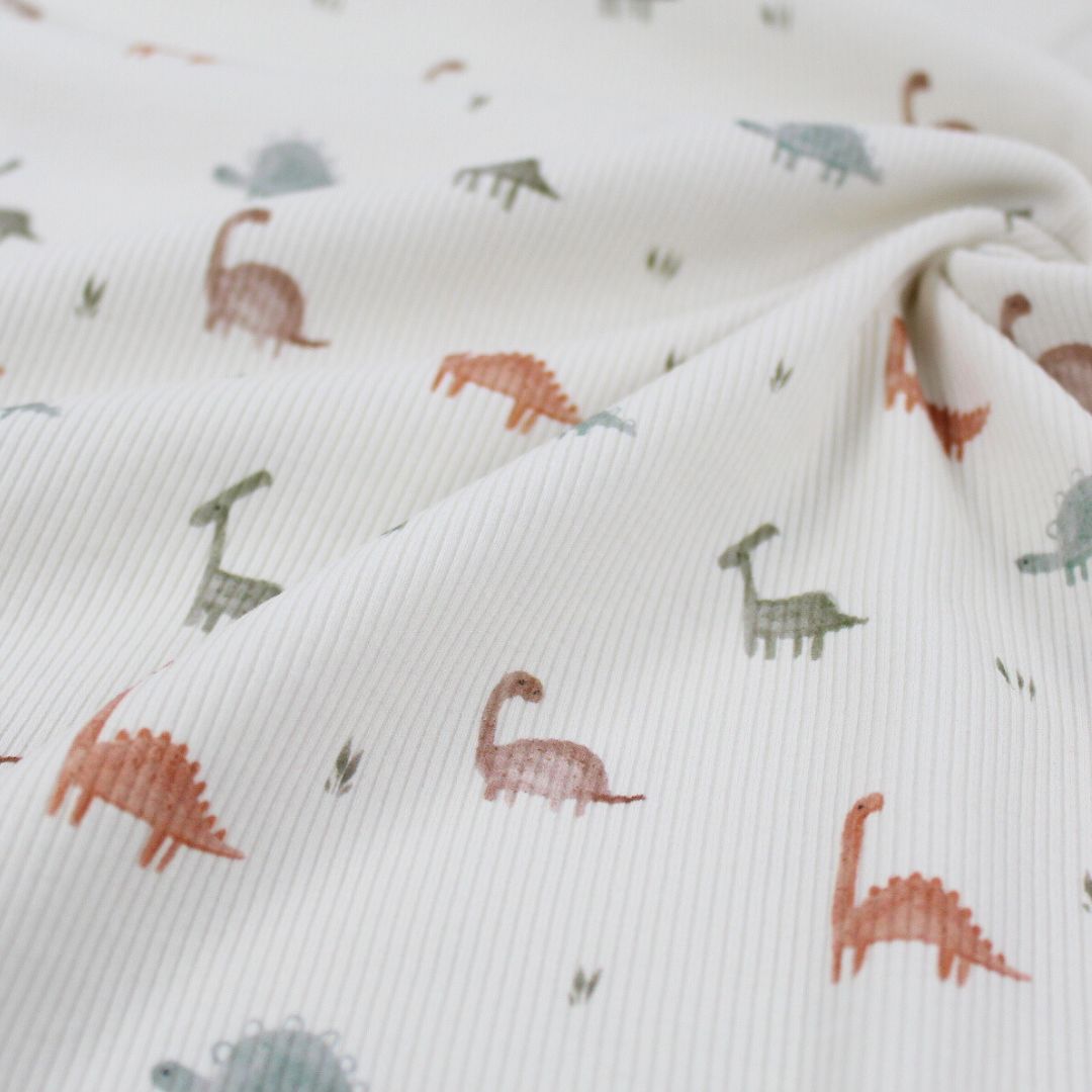 Weicher Rib Jersey Stoff mit bunten Dinosauriern auf ecrufarbenem Hintergrund – perfekt für bequeme Kinderkleidung.